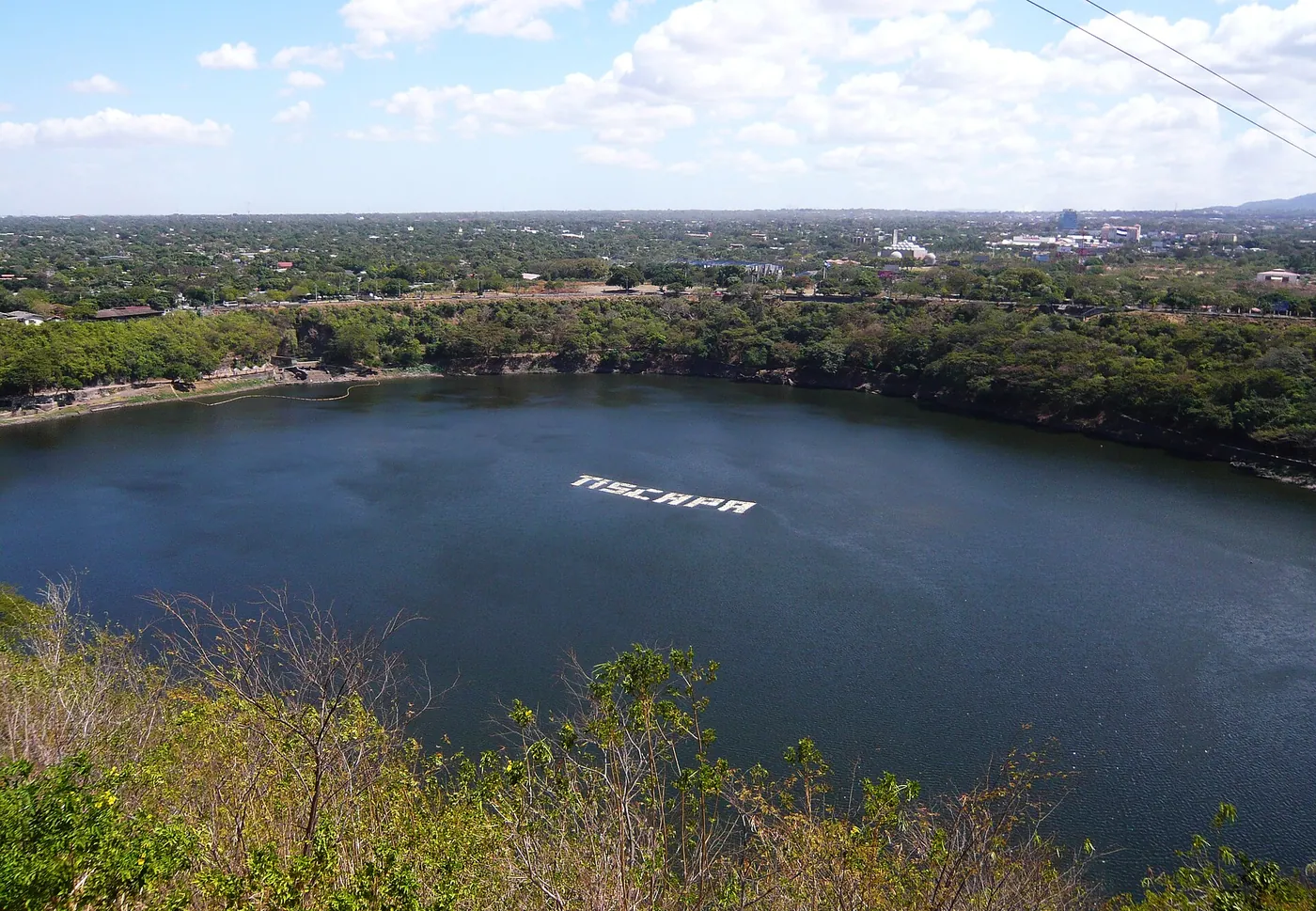 A round crater lagoon surrounded by trees with the city beyond